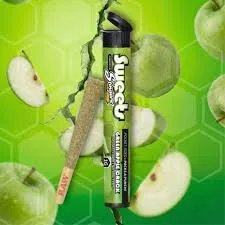 Black and green tube labeled Green Apple Crack beside a cone pre-roll, over a green apple background.