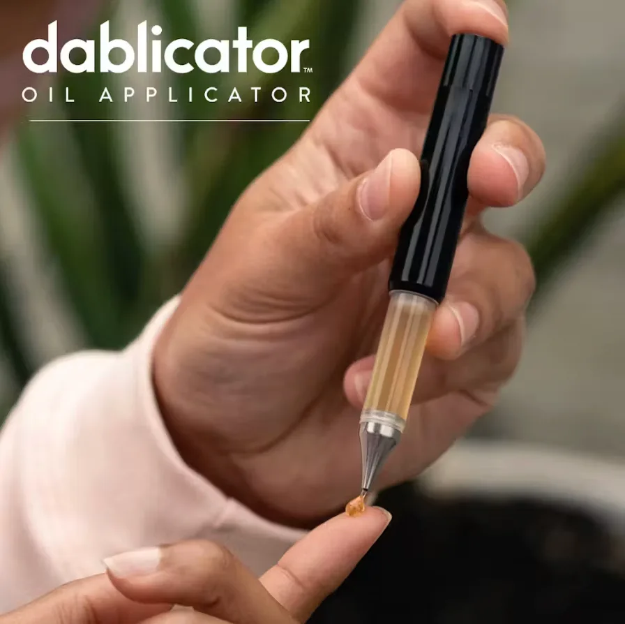 Hand holding a black Dablicator oil applicator with amber cannabis oil visible in its clear barrel, under the Dablicator Oil Applicator logo.