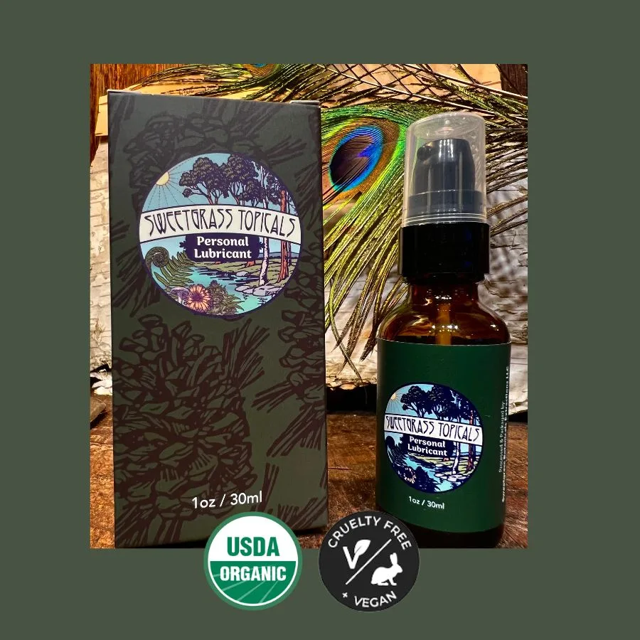 Dark gift box and amber glass pump bottle labeled Sweetgrass Topicals Personal Lubricant, 1 oz / 30 ml, beside a peacock feather.